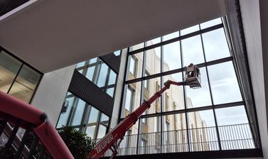Photo taken from inside the Life and Mind Building, towards a construction crane lifting a worker to work on the inside of the glass facade over the main entrance