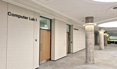 Interior view of computer lab entrances, off the lower atrium of the Life and Mind Building. White walls and ceiling, grey floor and pillars, and wood-effect doors.