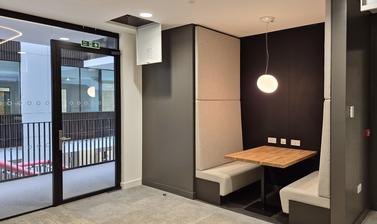 A meeting booth in the Life and Mind Building, with tall beige bench-seats, wooden table, egg-shaped light suspended from the white ceiling. Grey floor surrounding the booth and a view through glass doors into the central atrium