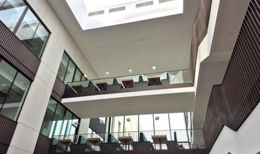 Interior view of the central atrium of the Life and Mind Building, looking up towards the upper floors and glass ceiling. White walls, black railings, and soft seating with tables in the distance.