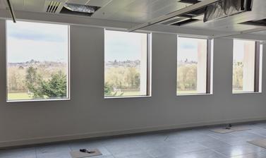View towards four rectangular glass windows from an upper floor space with flooring and ceiling still under construction in the Life and Mind Building; view towards Oxford spires, green space and trees