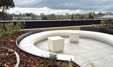 View of part of a cafe terrace on the upper floor of the Life and Mind Building on a cloudy day, looking towards Oxford's spires and trees; new planting and curved stone seating in the foreground