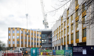 View of emerging entrance to Life and Mind Building (exterior), on a cloudy November day, with a construction crane in the background and security fencing in the foreground