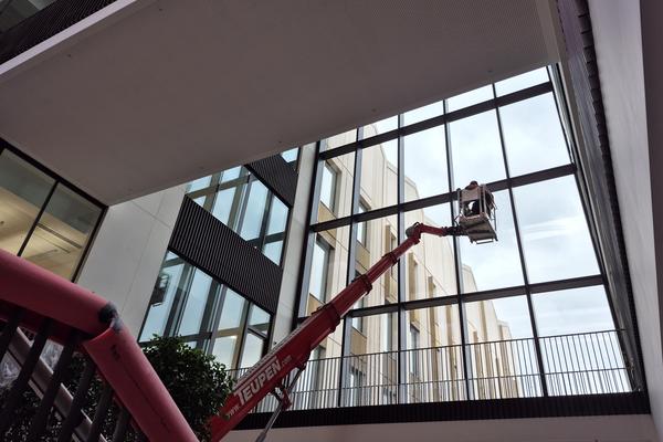 Photo taken from inside the Life and Mind Building, towards a construction crane lifting a worker to work on the inside of the glass facade over the main entrance