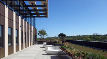 View of one side of The Life and Mind Building from the upper floor, towards a part paved, part planted terrace, with blue sky and distant greenery in the background